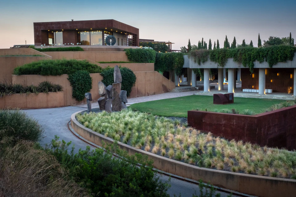Modern building with terraces, surrounded by greenery. Sculptures and manicured garden with pathway in the foreground. Twilight sky.
