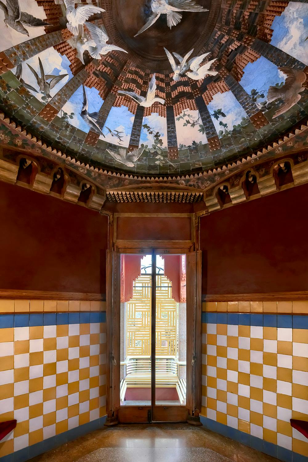 Elaborate ceiling with bird mural, ornate door with geometric pattern, and checkered yellow and white tiled walls.