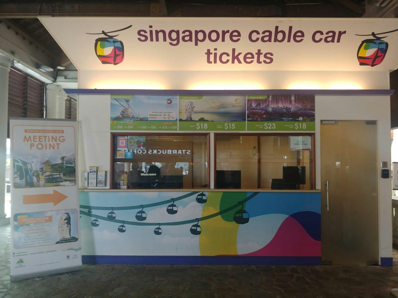 Comptoir de vente de billets pour le Singapore Cable Car, avec des panneaux de prix et des affiches promotionnelles, avec un panneau de bienvenue et une peinture murale colorée en dessous.