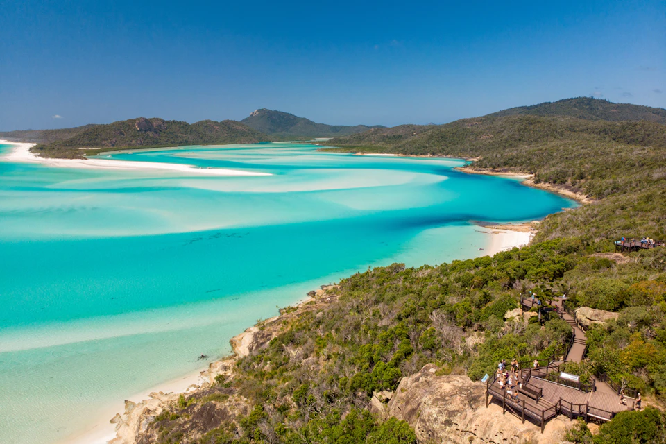 A scenic view overlooking a turquoise lagoon with a wooden viewing platform and people on the right, surrounded by lush greenery.