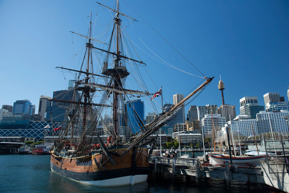Tall wooden sailing ship docked at a marina with city skyscrapers and a tower in the background under a clear blue sky.
