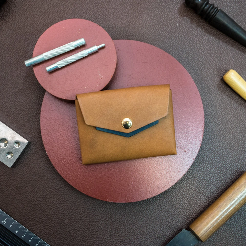 A brown leather wallet with a button closure on a red circular surface, surrounded by crafting tools on a dark background.