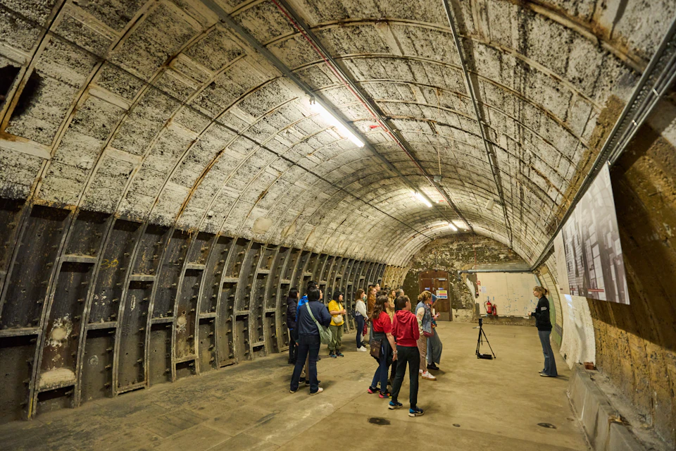A group of people standing and observing an arched underground tunnel with a weathered ceiling and walls.