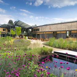 Modern byggnad med skylten "RHS Hilltop The Home of Gardening Science", omgiven av färgglada blomsterträdgårdar och uteserveringar.