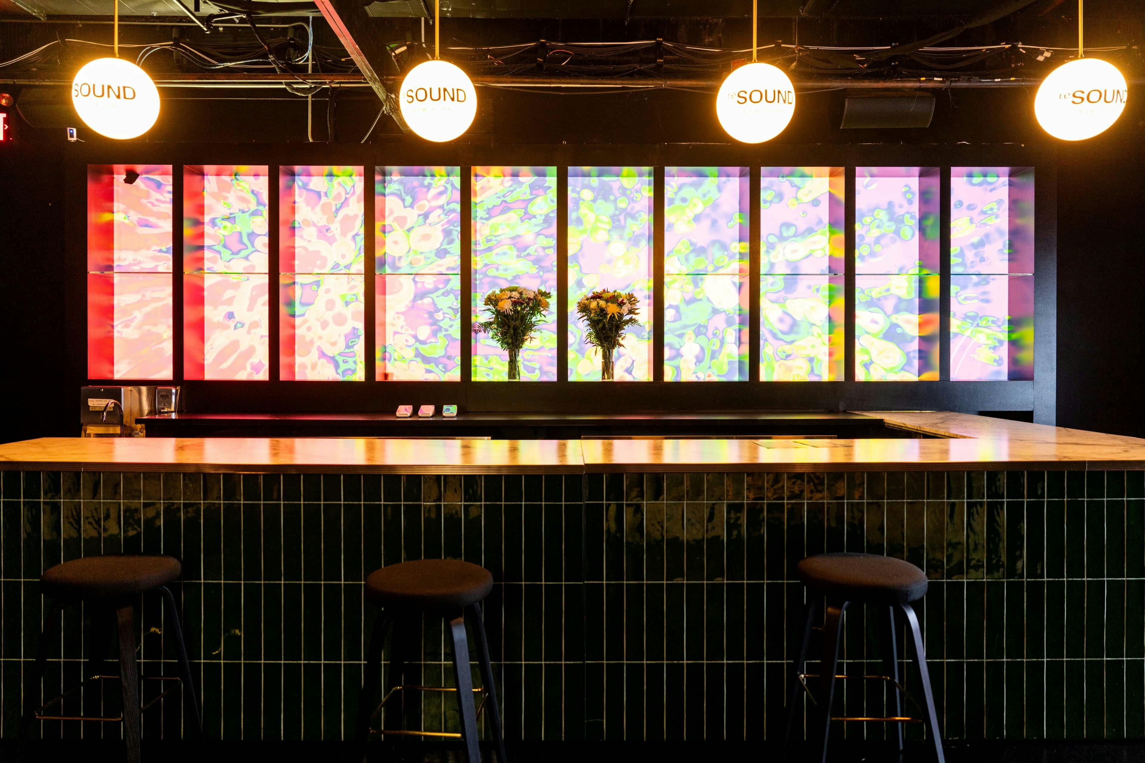 Bar counter with stools, two vases of flowers, colorful geometric wall panels, and two spherical lights labeled "SOUND."