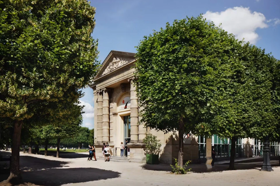 A classical-style building with columns and pediment, surrounded by trees, with people walking nearby under a blue sky.