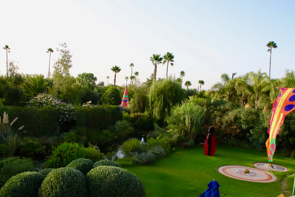 A lush garden with various plants, sculptures, a small pond, and palm trees under a clear sky.