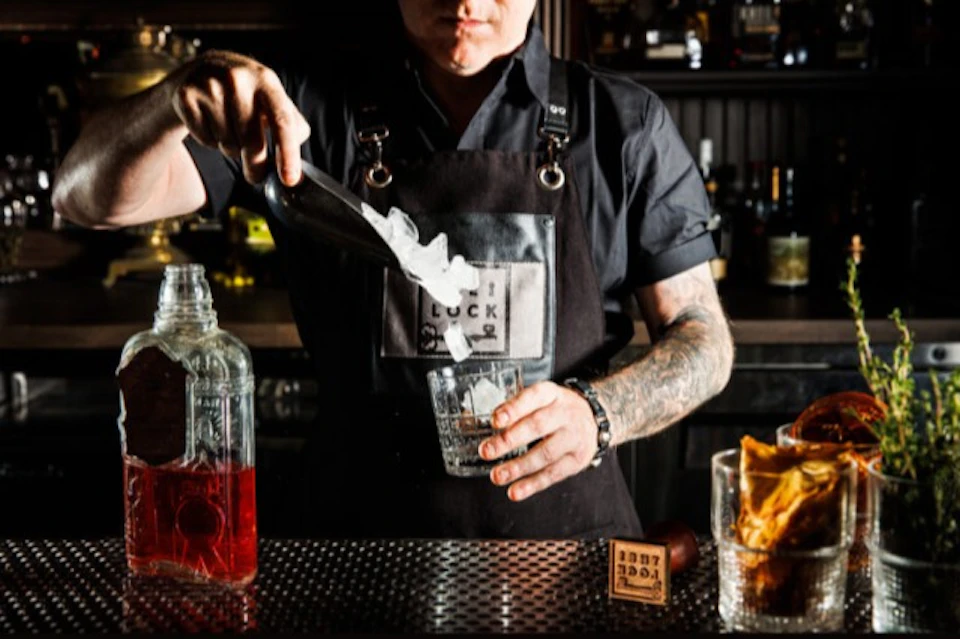A bartender in a black uniform adds ice to a glass at a bar, with bottles and oranges visible in the background.