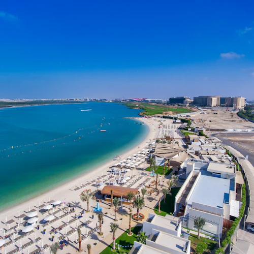 Luftaufnahme eines Strandresorts mit zahlreichen weißen Liegestühlen und Sonnenschirmen entlang des Ufers. Angrenzend befinden sich moderne Gebäude und eine große blaue Bucht.