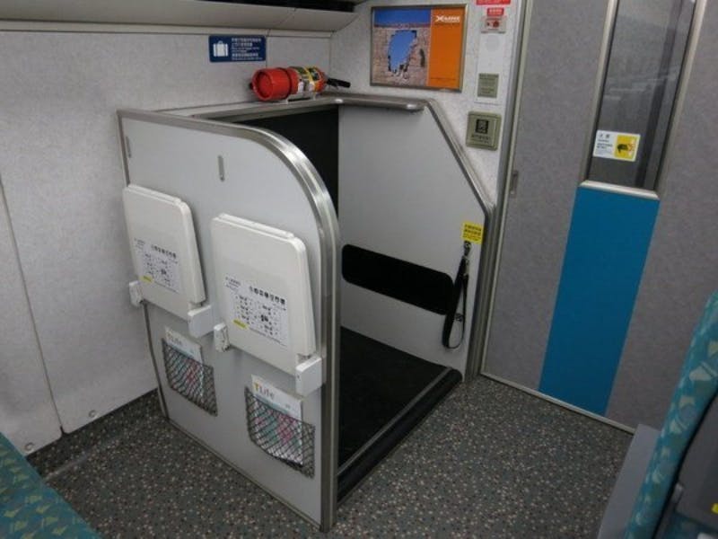 Κλειστός χώρος με πτυσσόμενα καθίσματα, πυροσβεστήρα και ενημερωτικές αφίσες σε έναν τοίχο, πιθανώς σε ένα τρένο.