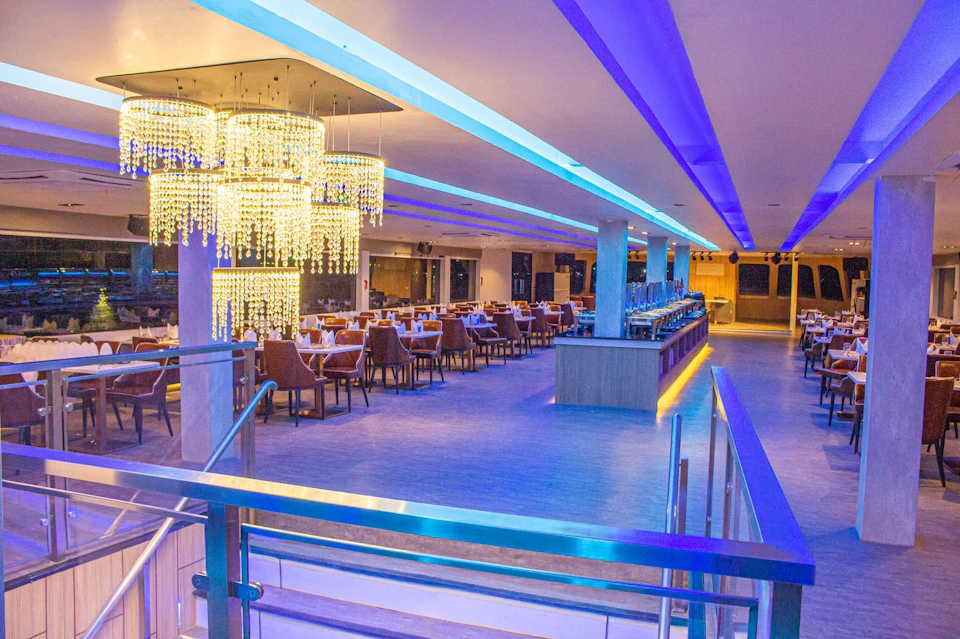 A modern restaurant with blue lighting, chandelier, neatly arranged tables, and a buffet counter. Stairs with glass railing in the foreground.