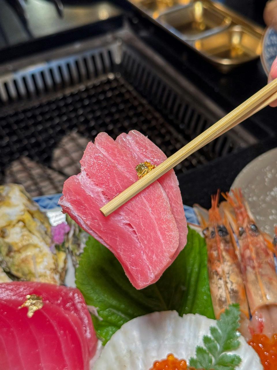 A piece of sashimi garnished with gold leaf held by chopsticks over a plate with seafood.