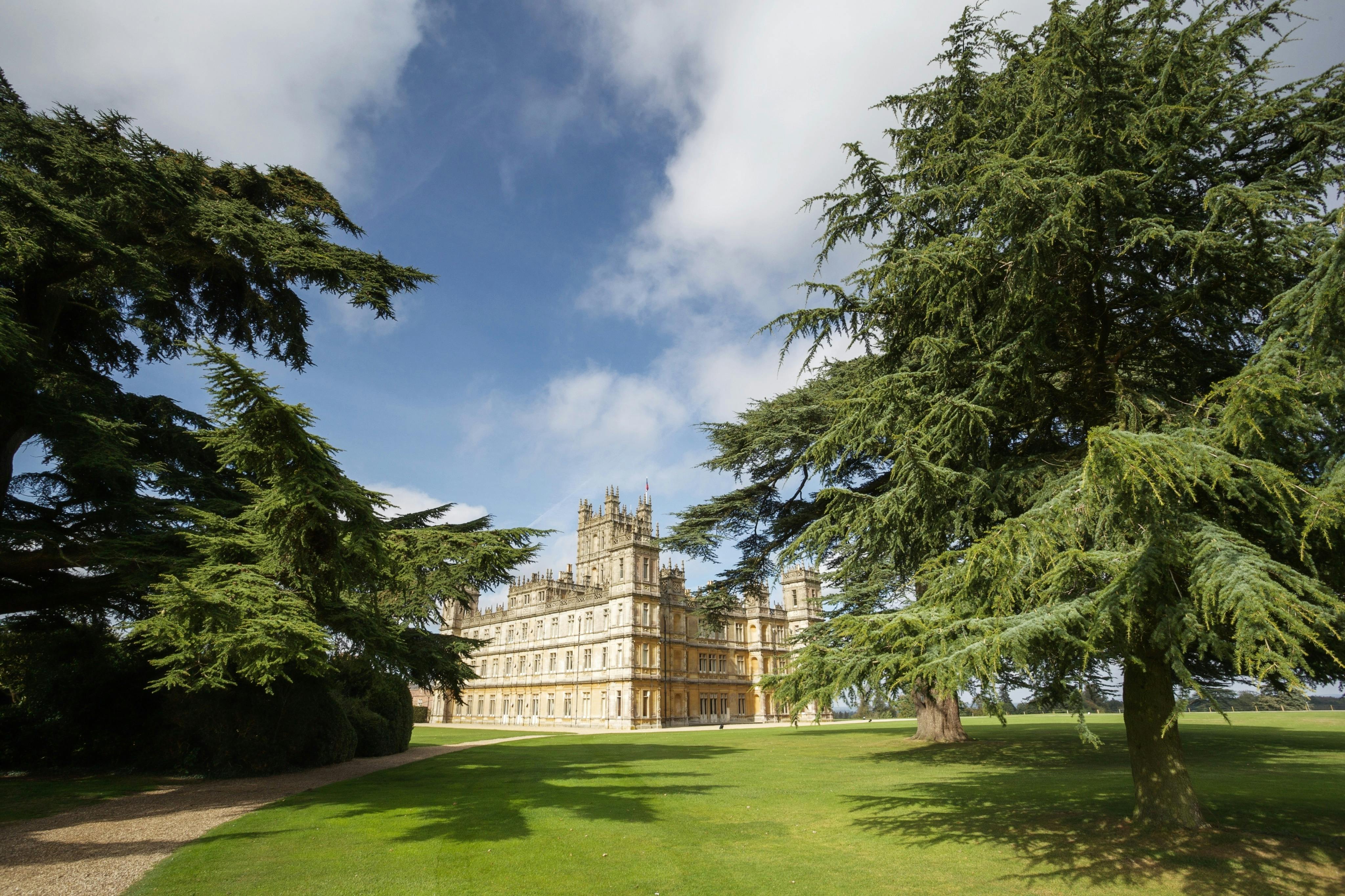 Een historisch, kasteelachtig gebouw met torentjes, omringd door groene gazons en hoge bomen onder een gedeeltelijk bewolkte hemel.