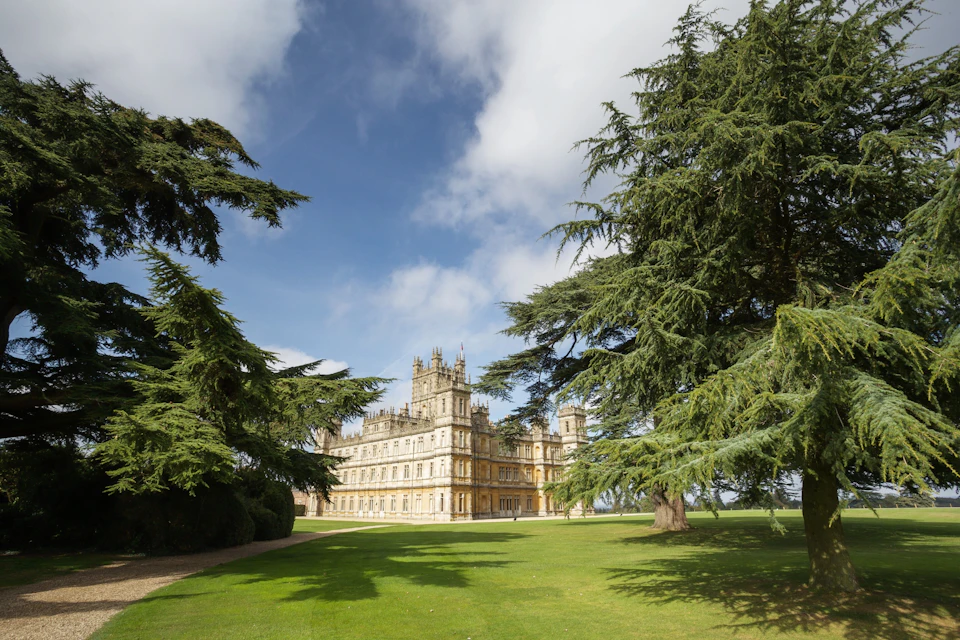 A historic, castle-like building with turrets, surrounded by green lawns and tall trees under a partly cloudy sky.