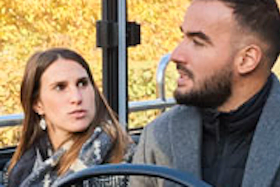 People seated inside a glass-roofed tour bus, looking at buildings and scenery outside. Bright and clear day.