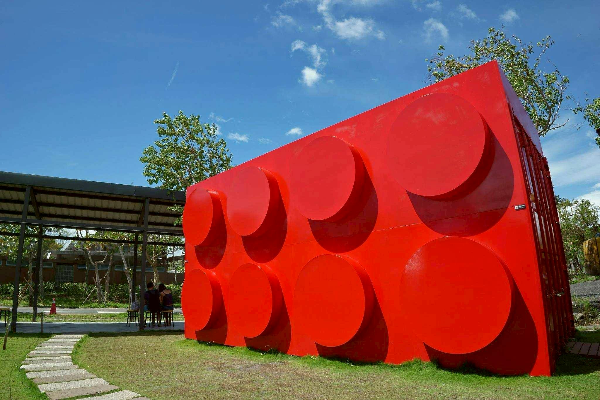 A vibrant red wall with circular patterns under a blue sky, next to a shaded structure and grass lawn.