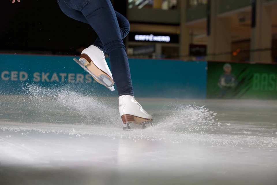 A person in white ice skates performing a move on an indoor ice rink, with water spraying from the ice.