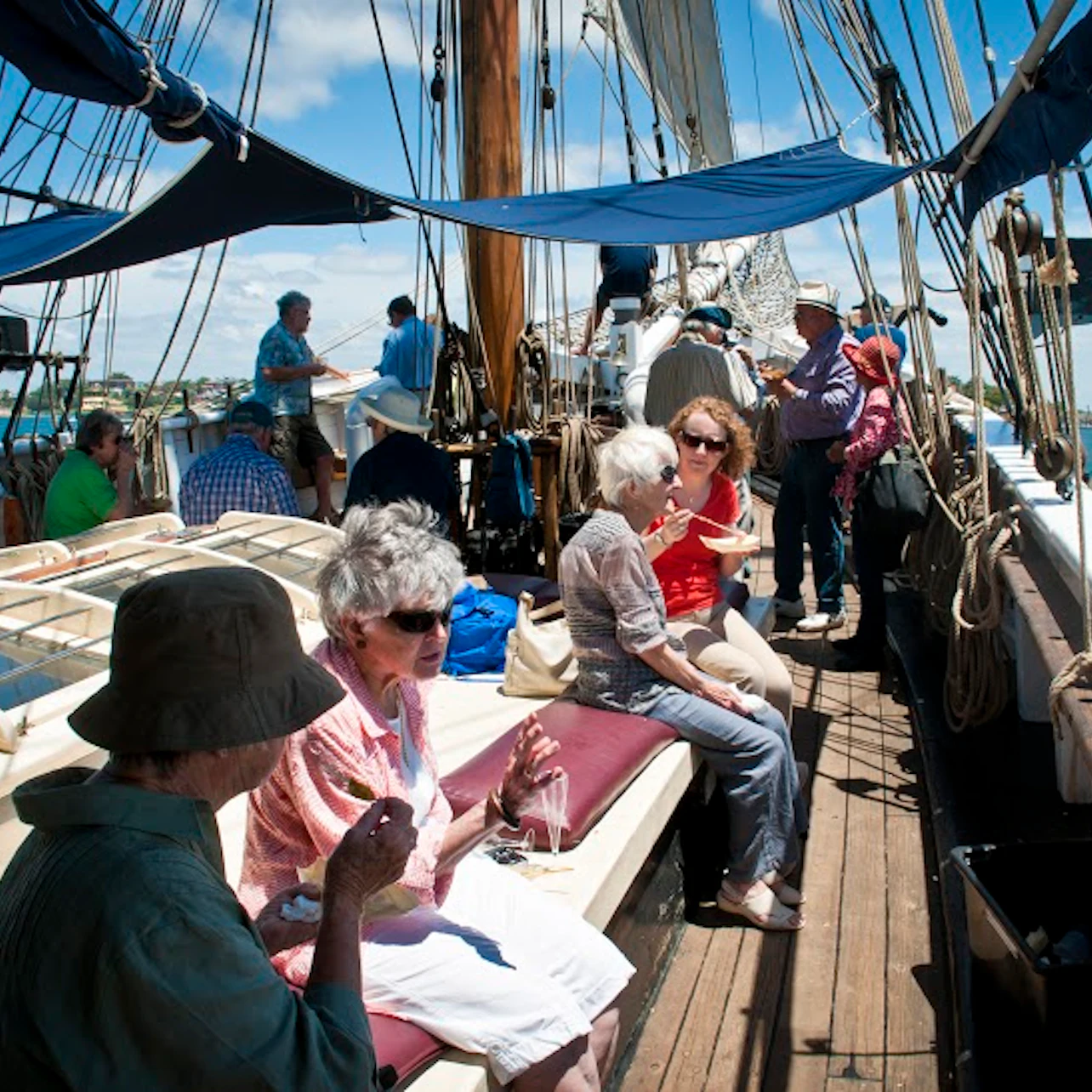People conversing and eating on the deck of a sailboat, surrounded by ropes, masts, and sails under a partly cloudy sky.