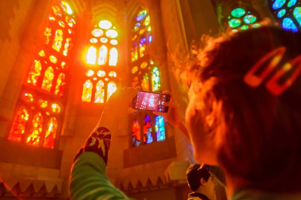 A person photographs colorful stained glass windows inside a cathedral, with sunlight streaming through.