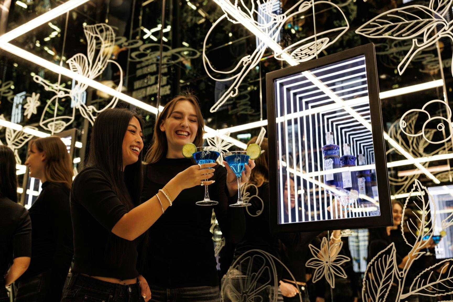两位女士微笑着吐司，在反光的霓虹灯背景下享用蓝色鸡尾酒，背景装饰着花卉图案。