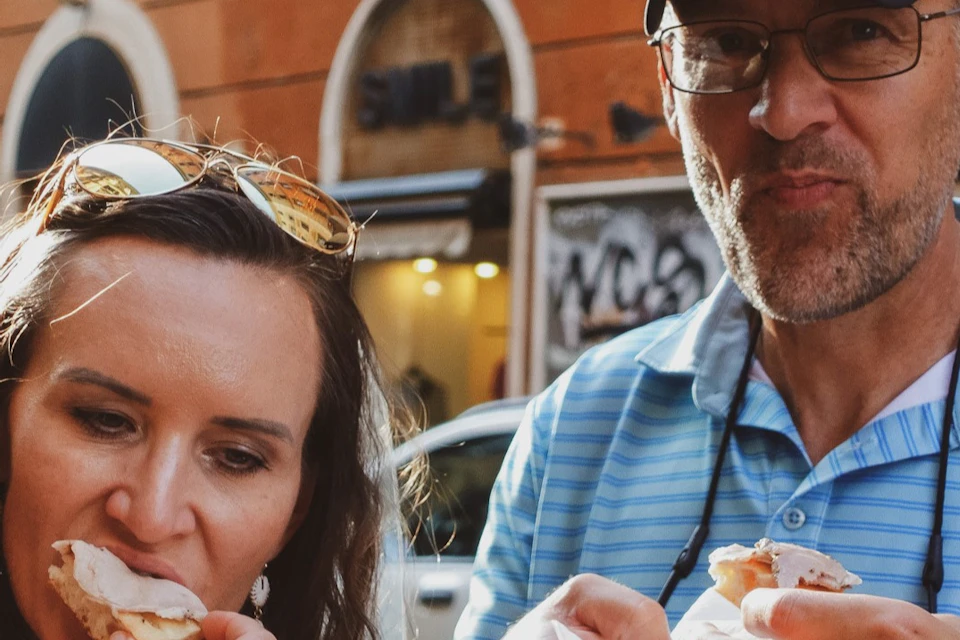 Two people enjoying food outdoors in an urban setting, with a brick building and arched windows in the background.