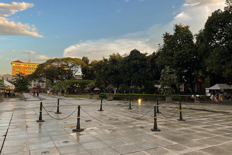 A paved plaza with chain barriers, surrounded by trees and greenery under a partly cloudy sky during sunset.