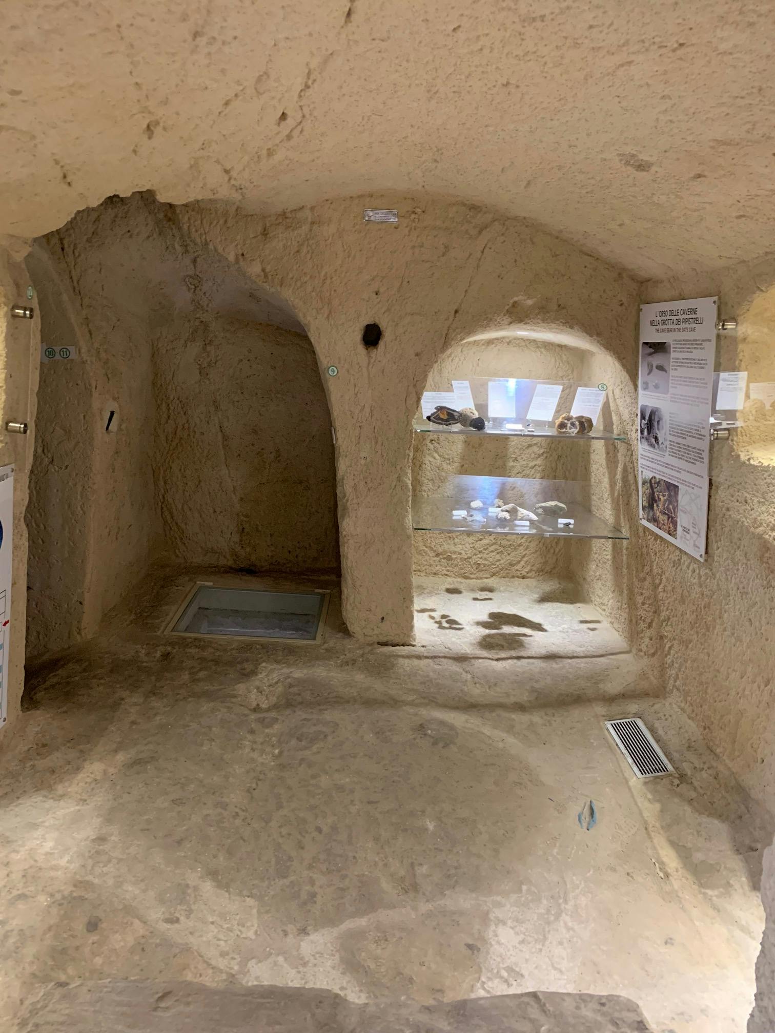 Mostra di una grotta sotterranea con teschi e ossa esposti su scaffali di vetro, pannelli informativi sulle pareti e una piccola apertura.