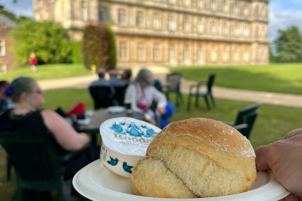 A plate with a bread roll and a container of spread is held in the foreground, with people seated outdoors near a large building.