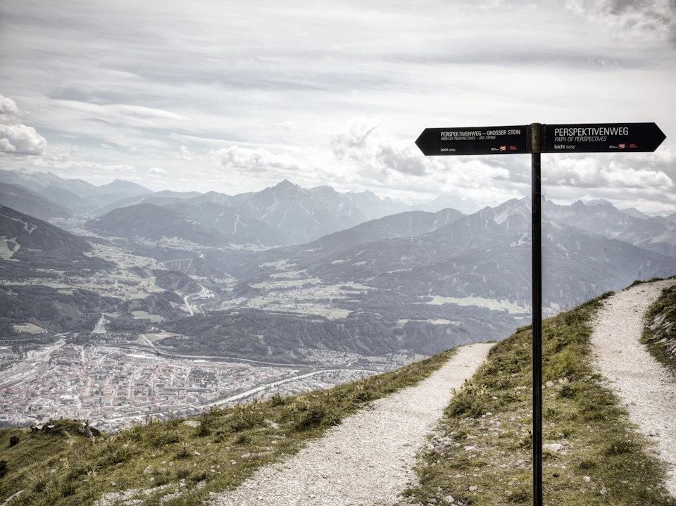 Mountain path with a signpost pointing to different trails, overlooking a valley and town with distant mountain ranges in the background.