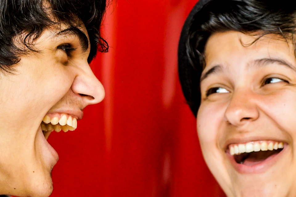 Two people laughing and holding drinks in front of a large Coca-Cola sign. One wears a hat and a camera strap.