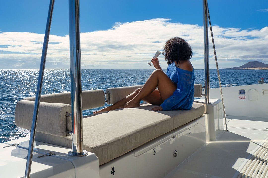 파란색 덮개를 쓴 곱슬머리를 한 사람이 보트의 쿠션이 있는 좌석에 앉아 음료를 들고 부분적으로 흐린 하늘 아래에서 바다를 바라보고 있습니다.