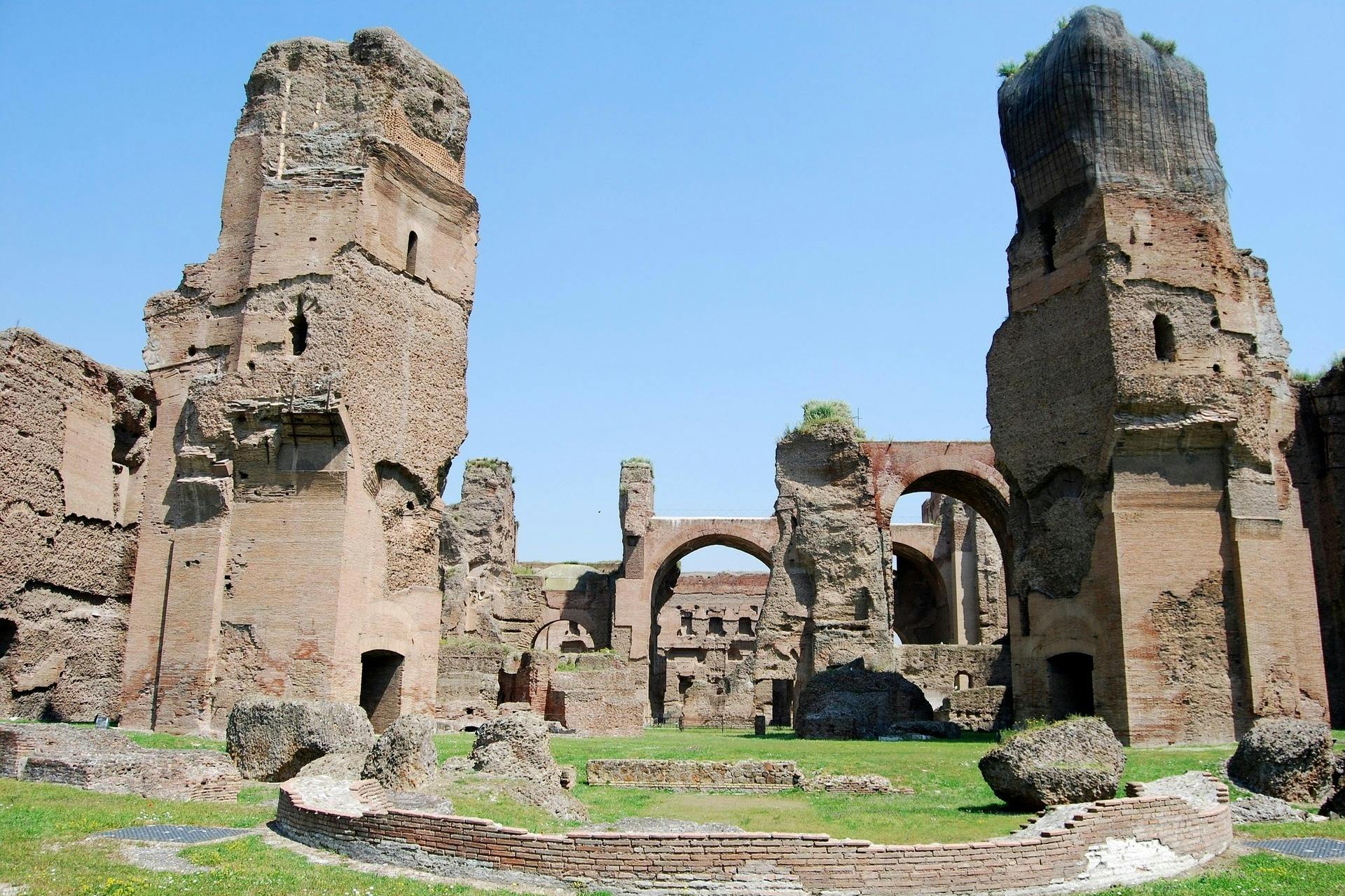 Ancient stone ruins with large arches and tall, weathered walls against a clear blue sky. Grass and small stone structures in the foreground.