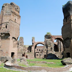 Ancient stone ruins with large arches and tall, weathered walls against a clear blue sky. Grass and small stone structures in the foreground.