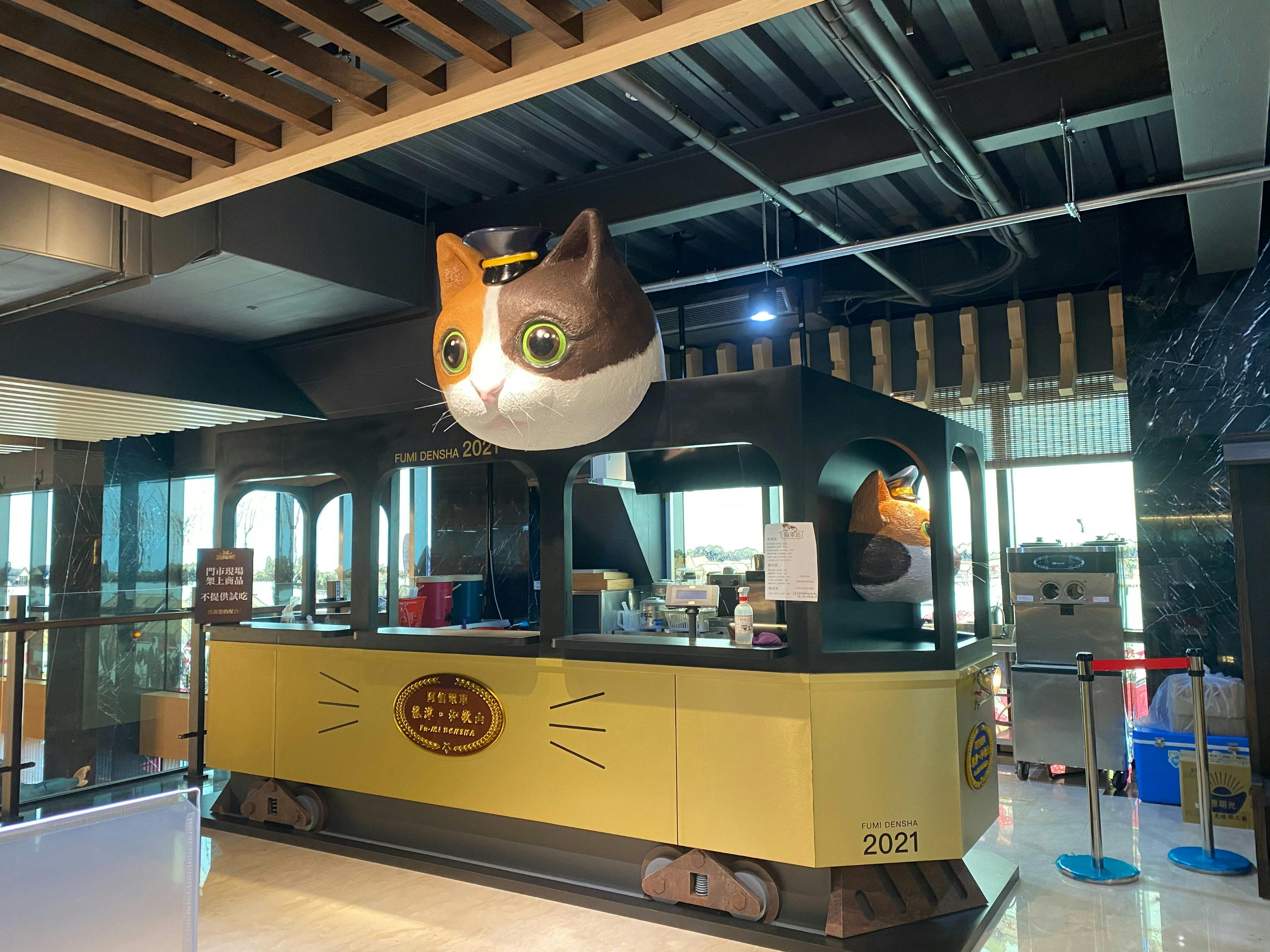 A food stall designed as a train car with a large cat head on the roof, located indoors under a wooden ceiling structure.