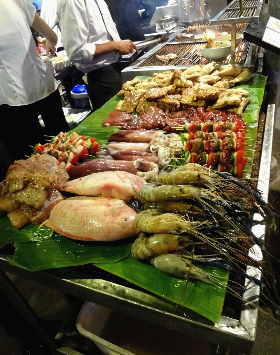 A variety of seafood, meats, and vegetable skewers are displayed on green leaves at a food market stall. Two people are grilling in the background.