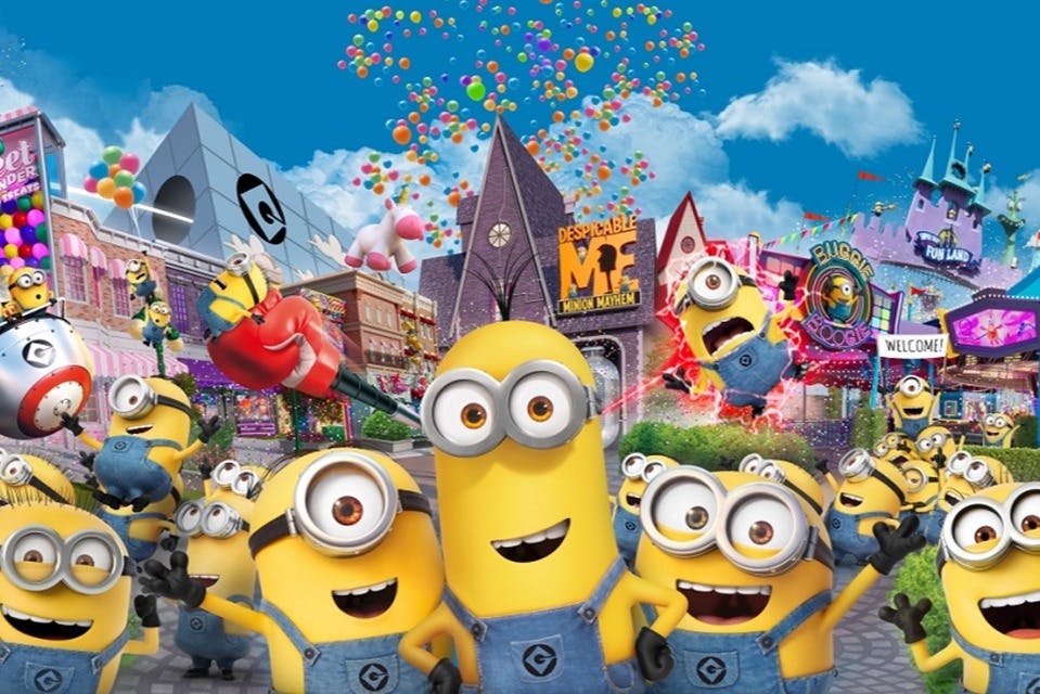 Skupina žlutých kreslených postaviček v montérkách v barevném prostředí zábavního parku s balónky a modrou oblohou.