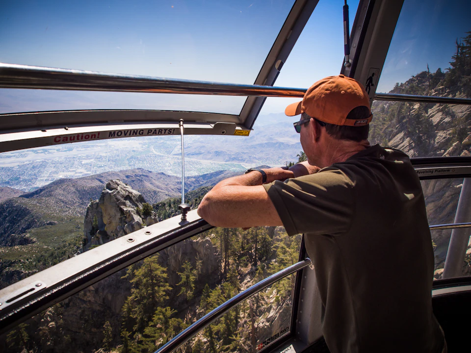 Man in an orange cap looking out a window of a cable car, overlooking a mountainous landscape with trees and rocky peaks.
