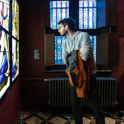 En man undersöker ett färgglatt glasmålningsfönster i ett svagt upplyst rum med mönstrade kakelplattor och fönsterväggar.