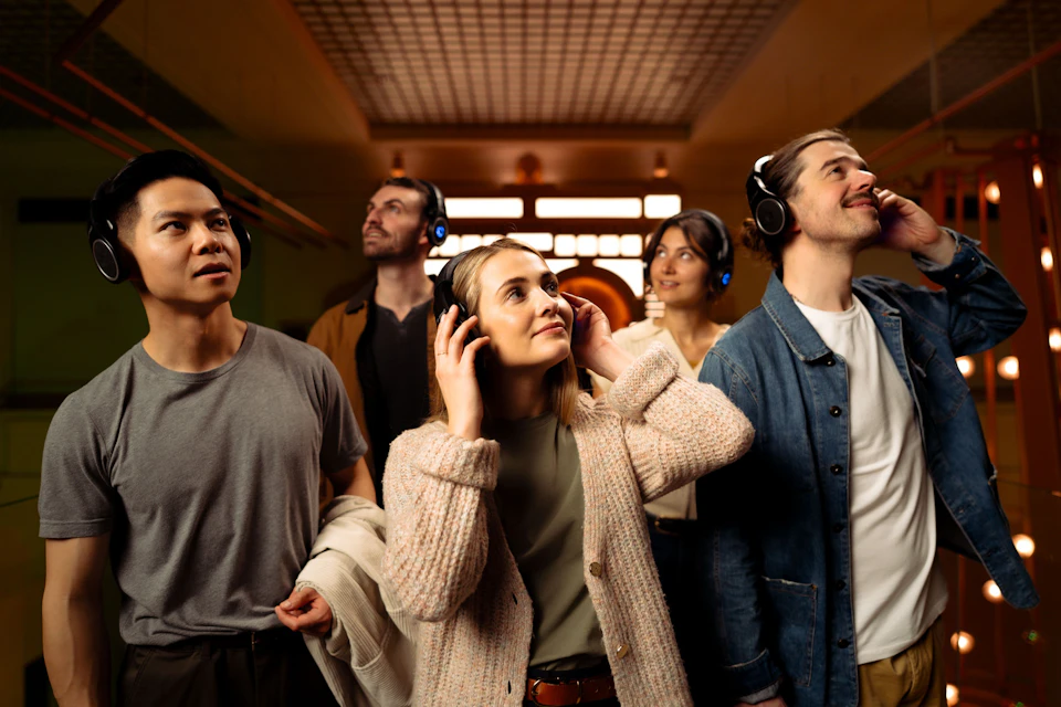 A group of four people wearing headphones, standing in a warmly lit indoor space, looking up and appearing engaged.