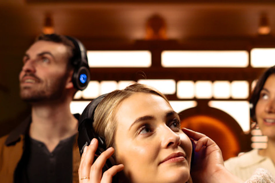 A group of four people wearing headphones, standing in a warmly lit indoor space, looking up and appearing engaged.