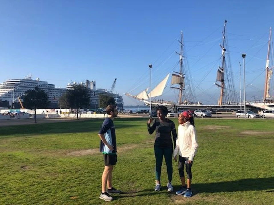 Three people standing on a grassy area near a harbor with a docked tall ship and cruise ship visible in the background.