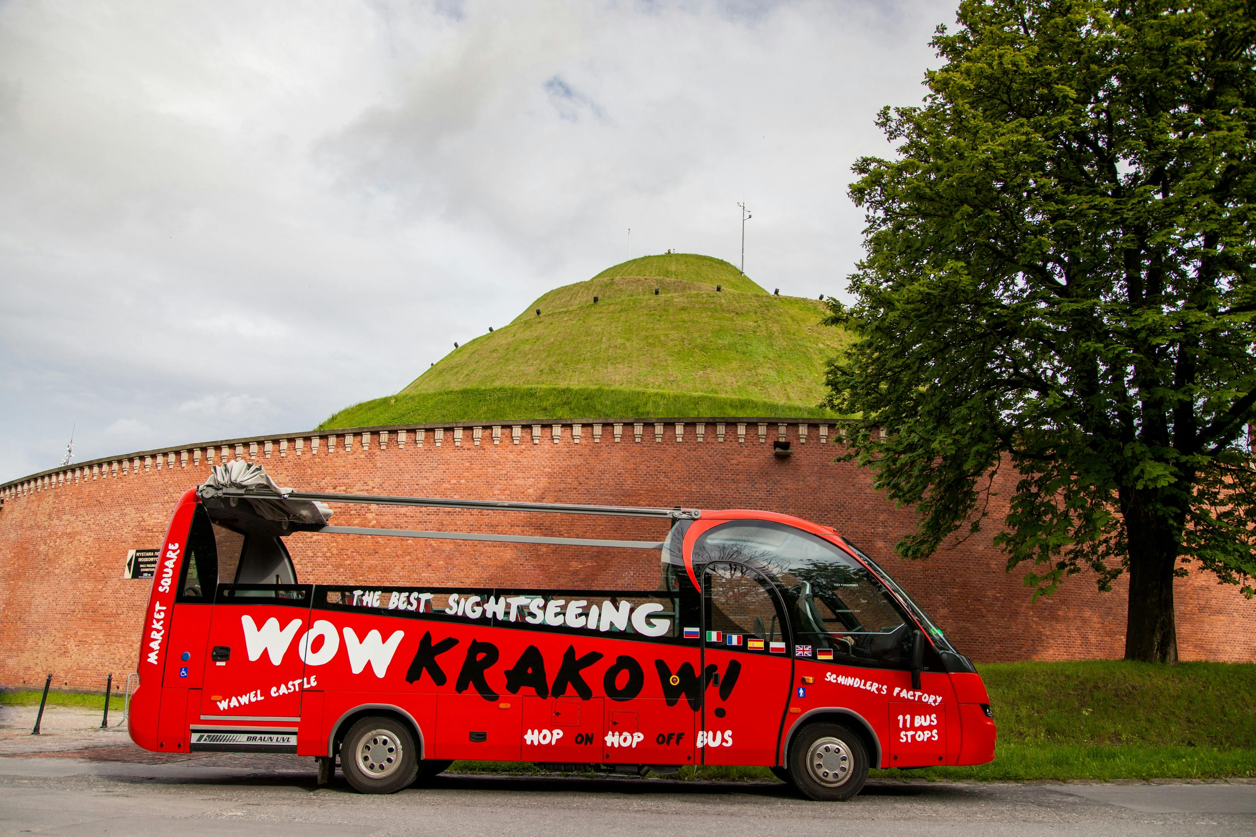Red sightseeing bus with “WOW KRAKOW!” parked in front of a grassy mound with a brick wall and trees.