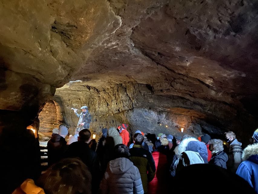 A group of people in winter clothing listen to a guide speaking and gesturing inside a dimly lit cave.