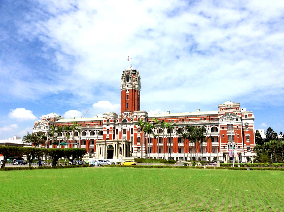 A grand red and white colonial-style building with a tall clock tower, surrounded by greenery, under a partly cloudy sky.