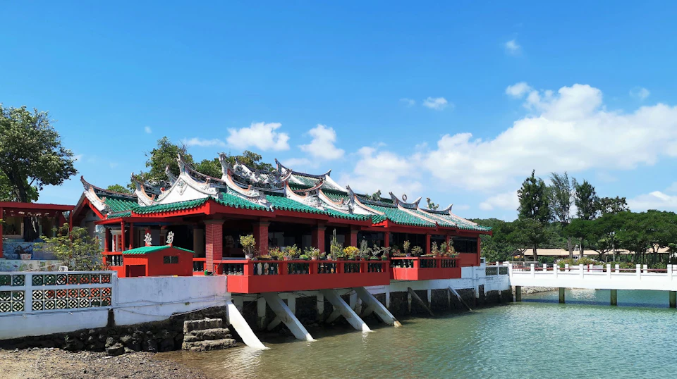 A red and white traditional Chinese building with ornate green roofs overhangs a body of water under a clear blue sky.