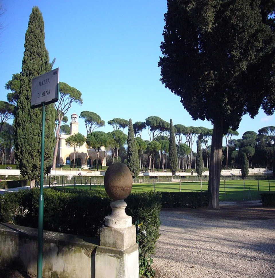 Park with trees, a gravel path, a sign reading "Piazza di Siena," and a stone structure in the foreground. Clear, sunny sky.