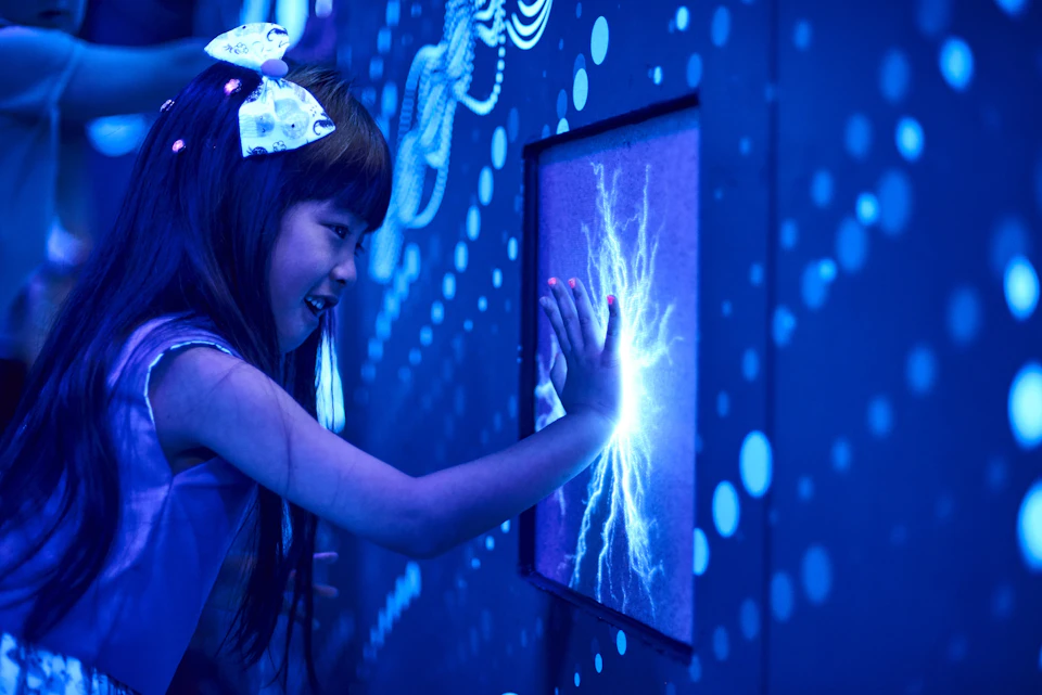 A girl touches a glowing blue screen showing electric-like patterns, surrounded by a dark, dotted, and illuminated wall.