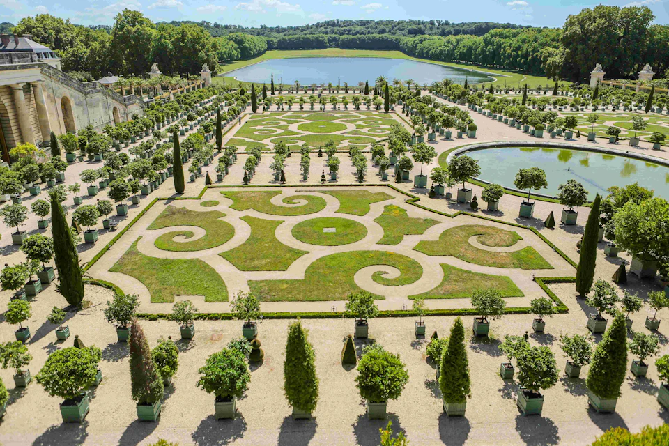 Intricately designed gardens with symmetrical patterns, hedges, and potted trees, overlooking a large lake surrounded by forest.
