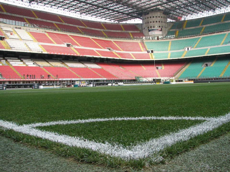 A view of an empty stadium with red and green seats, focusing on the corner of a soccer field marked with white lines.