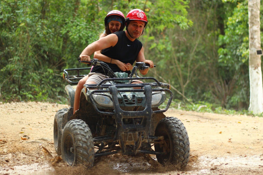 Deux personnes conduisant un VTT sur un sentier boueux dans la forêt, toutes deux portant un casque rouge et des vêtements décontractés, entourées de verdure.